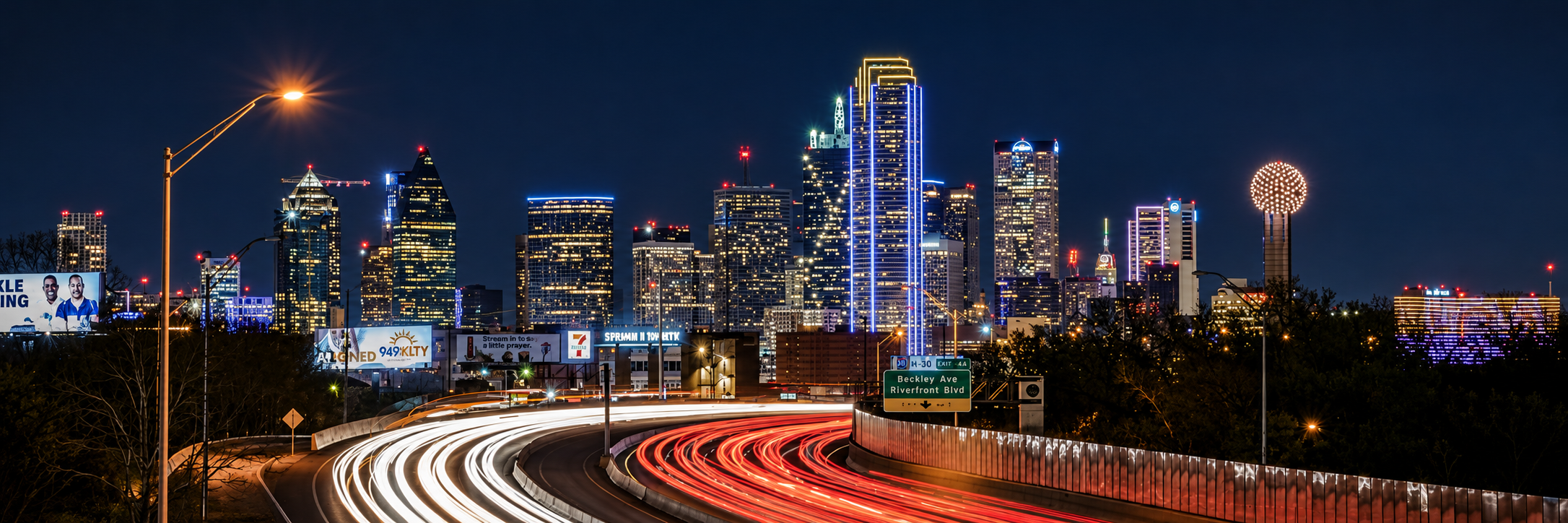 Dallas skyline at night, with illuminated towers above sweeping white and red freeway light trails.